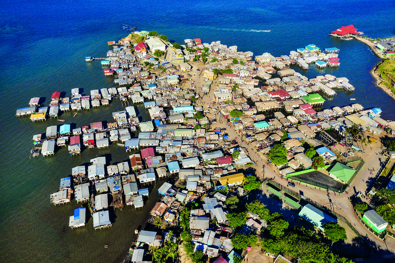 Fotografia. Vista aérea uma área urbana a beira-mar, ocupada por edificações horizontais. Destaque para uma superfície plana ocupada por ruas e casas situadas em braços  que avançam a superfície do mar.