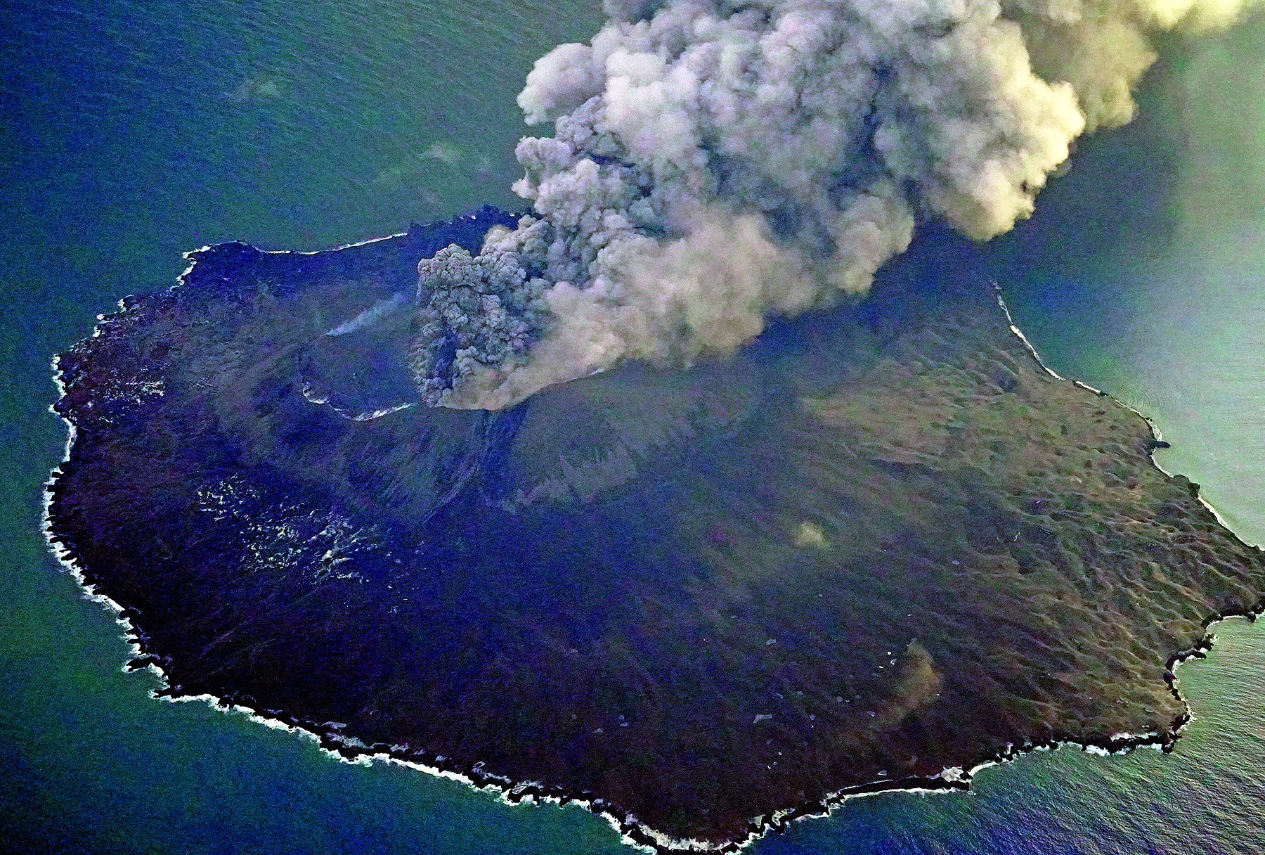 Fotografia. Vista aérea de uma ilha com um vulcão no centro, lançando fumaça densa na atmosfera. As vertentes do vulcão são cobertas por vegetação com folhagem escura. Ao redor da ilha, o mar.