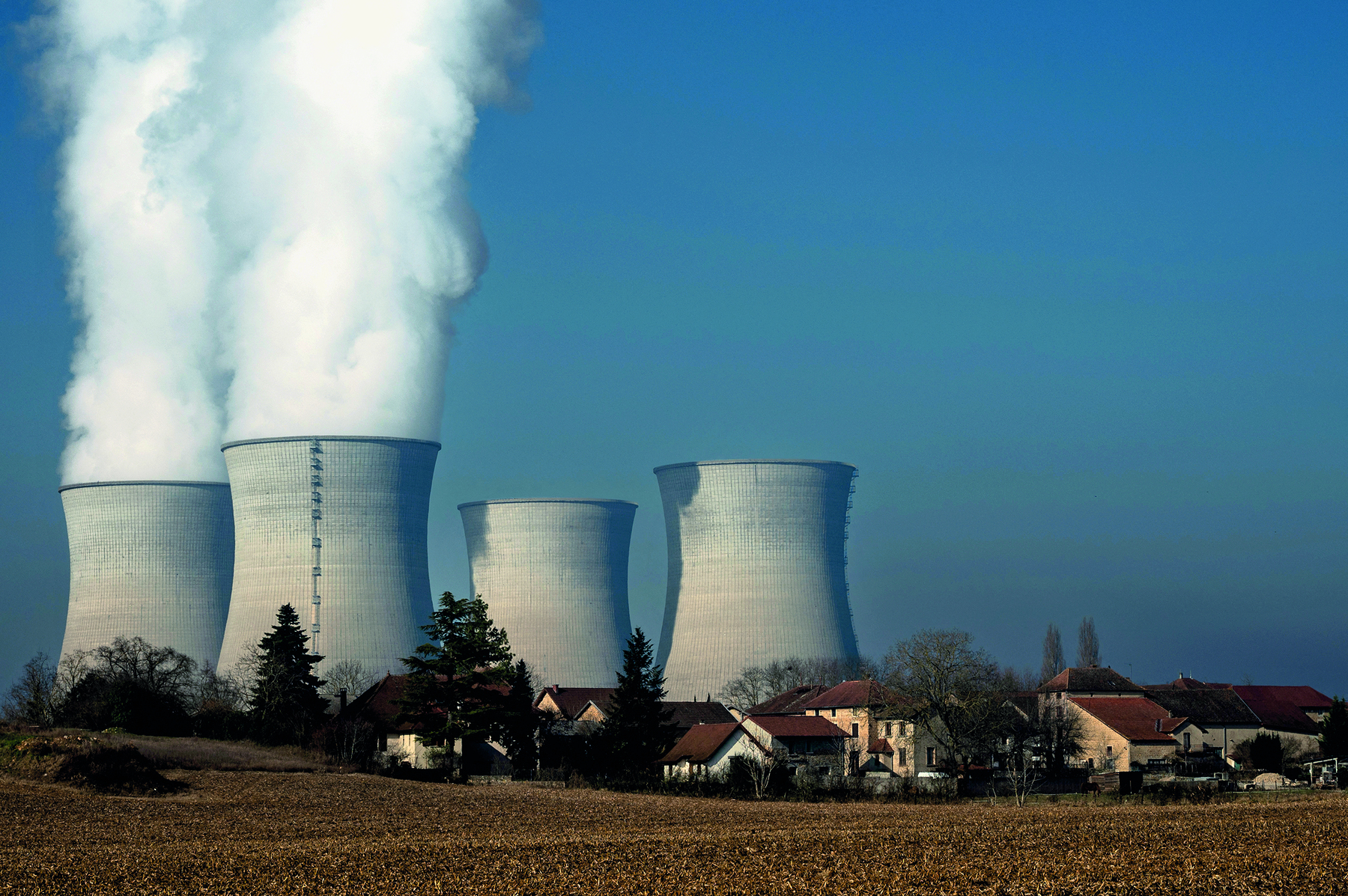 Fotografia. Destaque para uma paisagem composta pela presença de usina nuclear. No primeiro plano, superfície levemente inclinada coberta por vegetação rasteira. No segundo plano, presença de casas e árvores isoladas. No terceiro plano, quatro torres de resfriamento altas, brancas e de forma arredondada, sendo que duas lançam fumaça densa na atmosfera.