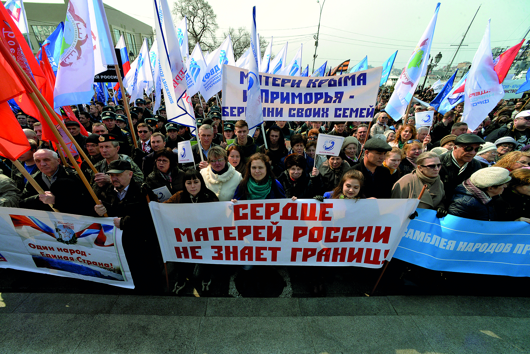 Fotografia. Manifestação de rua com multidão de pessoas em pé, segurando bandeiras, cartazes e faixas com textos no idioma estrangeiro.