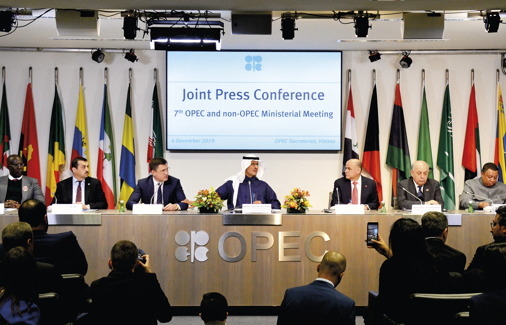 Fotografia. Vista para bancada de madeira escrito: OPEC. Atrás da bancada, há bandeiras nacionais nas laterais e um painel eletrônico no centro, com texto indicativo: Joint Press Conferente. 7th OPEC and non-OPEC Ministerial Meeting. Há homens sentados na bancada: no centro, um homem usando roupa azul e um lenço branco na cabeça; à direita e à esquerda dele, outros homens usando terno e gravata. Sobre a bancada, microfones, garrafas de água e vasos de flores. Na frente da bancada, diversas pessoas estão sentadas e assistem aos homens. Algumas delas seguram smartphones e câmeras fotográficas.