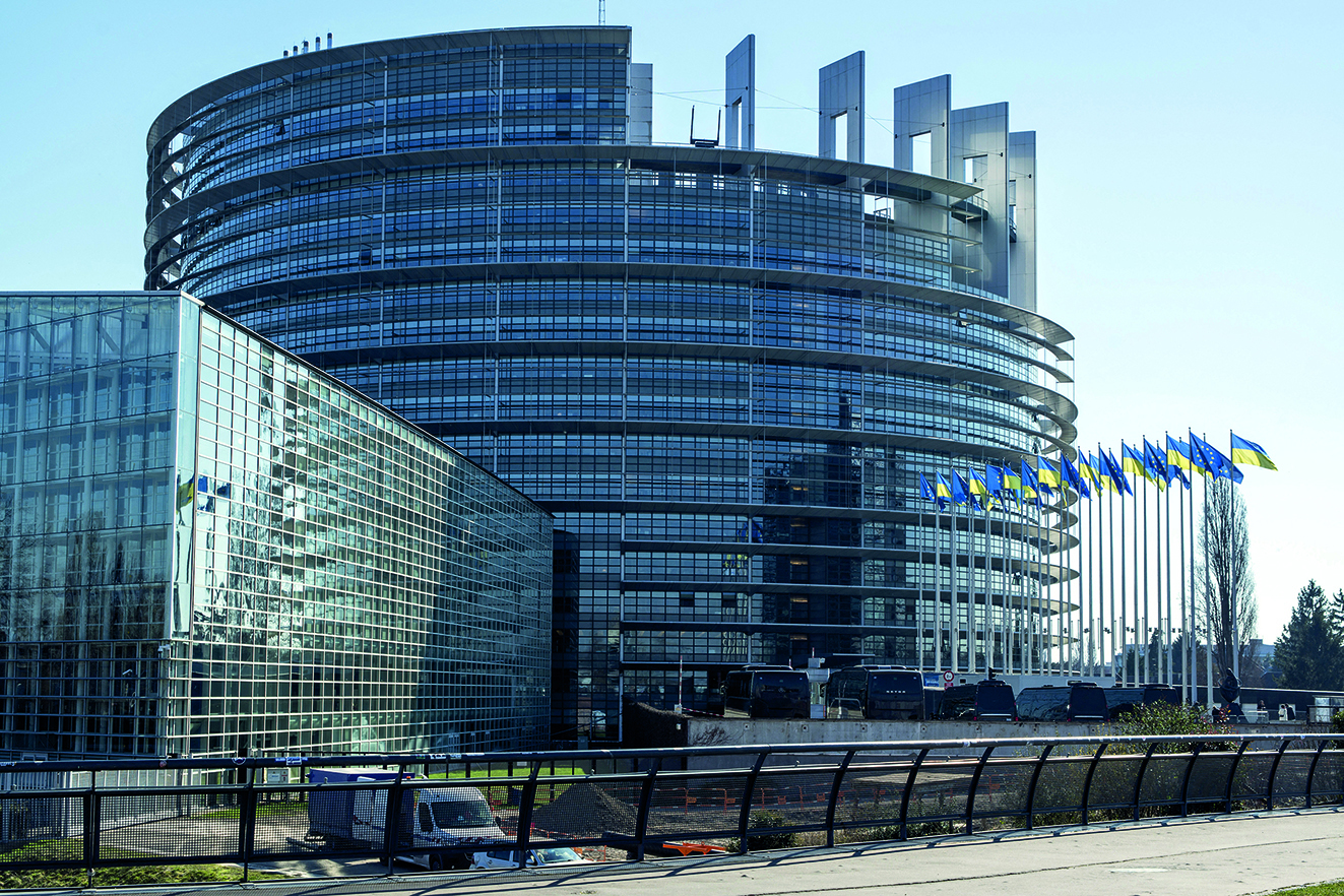 Fotografia. Vista para o edifício-sede do Parlamento Europeu. No primeiro plano, plataforma com parapeito de vidro. No segundo plano, um edifício redondo e espelhado no centro. À direita, há diversos mastros com bandeiras em tons de azul e amarelo. À esquerda, um edifício retangular e espelhado.