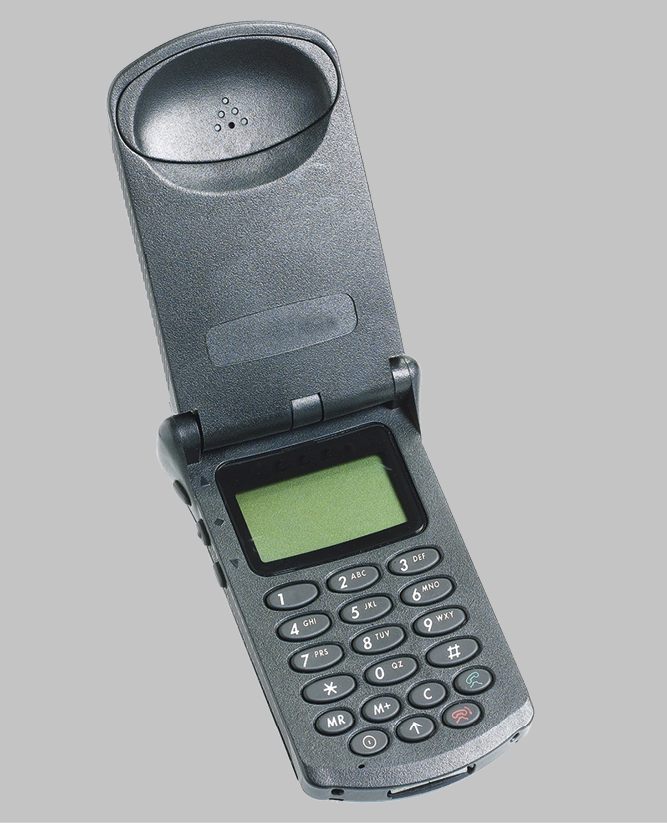 Fotografia. Destaque para um aparelho de celular antigo, pequeno, cinza e do tipo que abre e fecha. As teclas são ovais e a tela é pequena e verde.