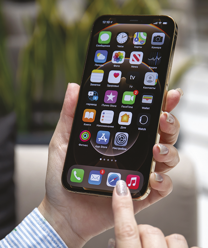 Fotografia. Um aparelho de smartphone moderno, retangular e preto, com tela grande e diversos símbolos de aplicativos. Uma mão inclinada e o dedo pressionando um dos aplicativos do aparelho.