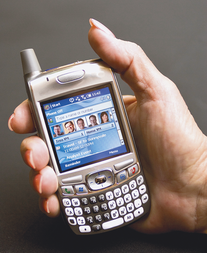 Fotografia. Destaque para um aparelho de celular antigo, retangular, com teclas pequenas, tela média azul com fotos de pessoas e uma pequena antena na parte superior.