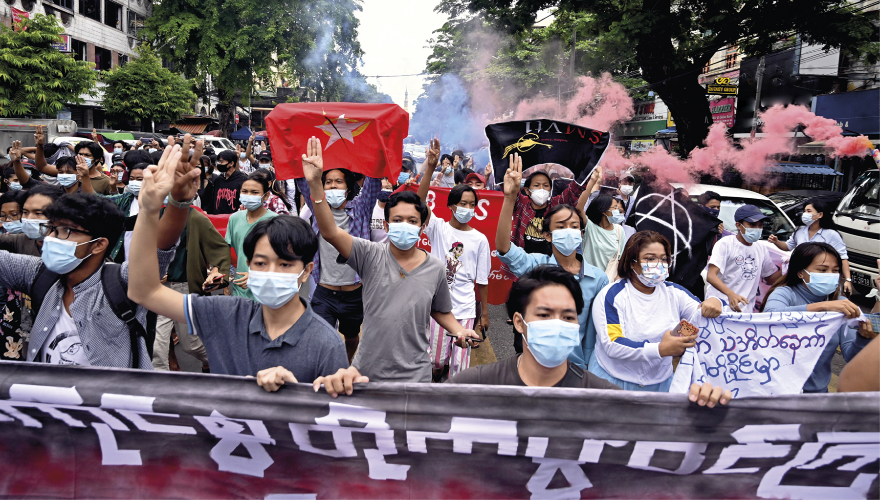 Fotografia. Destaque para uma manifestação de rua com diversas pessoas vestindo máscara de proteção facial. Algumas seguram faixas e bandeiras e outras erguem um dos braços. Ao fundo, há fumaça em tons de azul e vermelho, árvores e edificações.