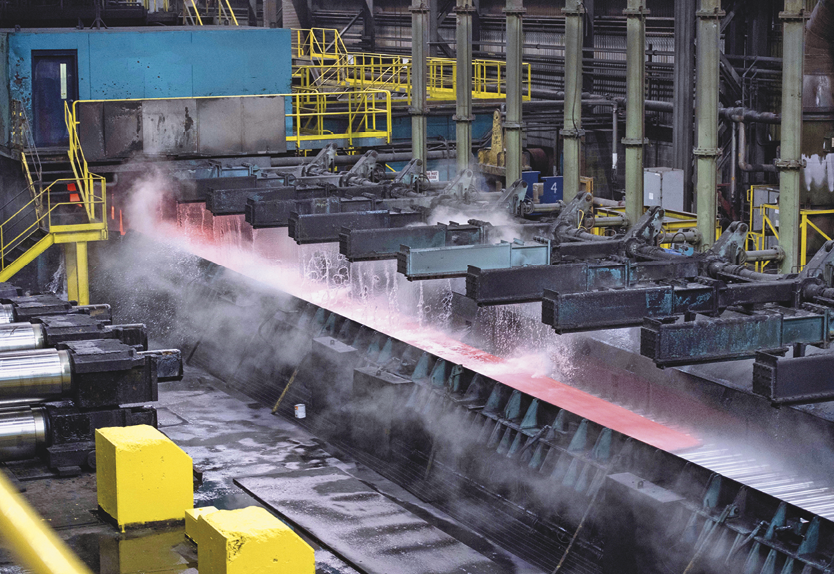 Fotografia. Vista para o ambiente interno de uma indústria. No centro, uma máquina com suportes paralelos jorrando água sobre aço quente. Ao redor há caixas, escadas e corrimões amarelos.