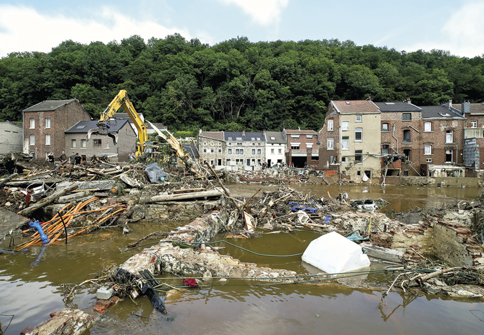 Fotografia. No primeiro plano, destaque para uma superfície plana inundada de água turva e lixo. Ao fundo, uma retroescavadeira limpa a superfície. No segundo plano, diversas casas e edifícios baixos com muitas janelas. No terceiro plano, floresta densa e árvores de grande porte com folhas verdes.