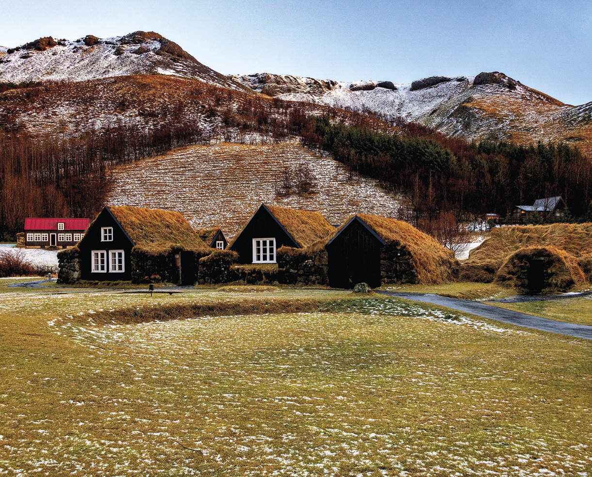 Fotografia. No primeiro plano, superfície plana, coberta por gramíneas. No segundo plano, pequeno curso d’água e três casas com revestimento escuro, janelas brancas e telhados de turfas em tons de verde e marrom. No terceiro plano, árvores altas e sem folhas e montanhas cobertas de neve.