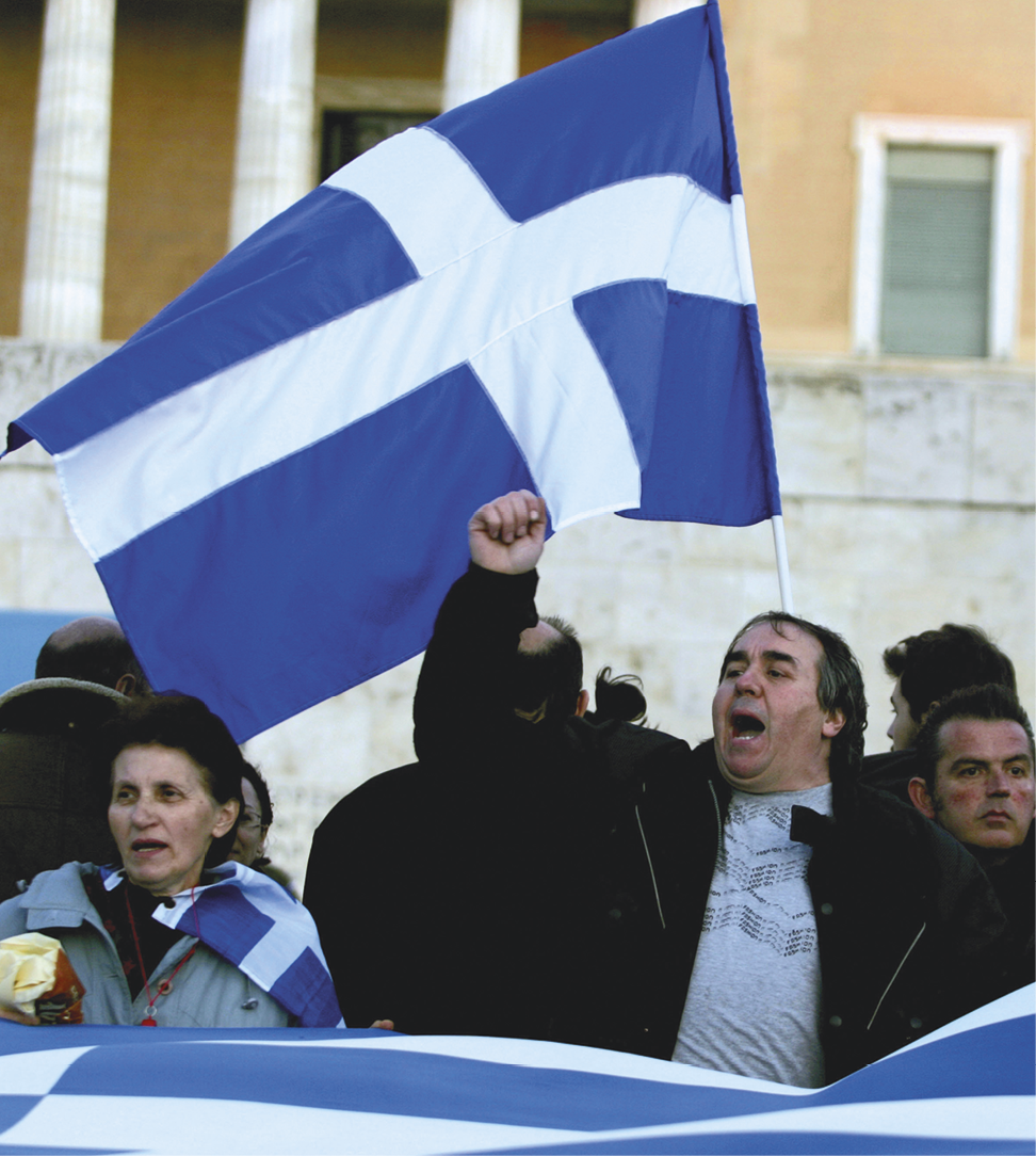 Fotografia. No primeiro plano, manifestação de algumas pessoas, com destaque para um homem de agasalho, mão direita erguida e boca aberta. Na frente e atrás das pessoas, a bandeira da Grécia, azul e branca.