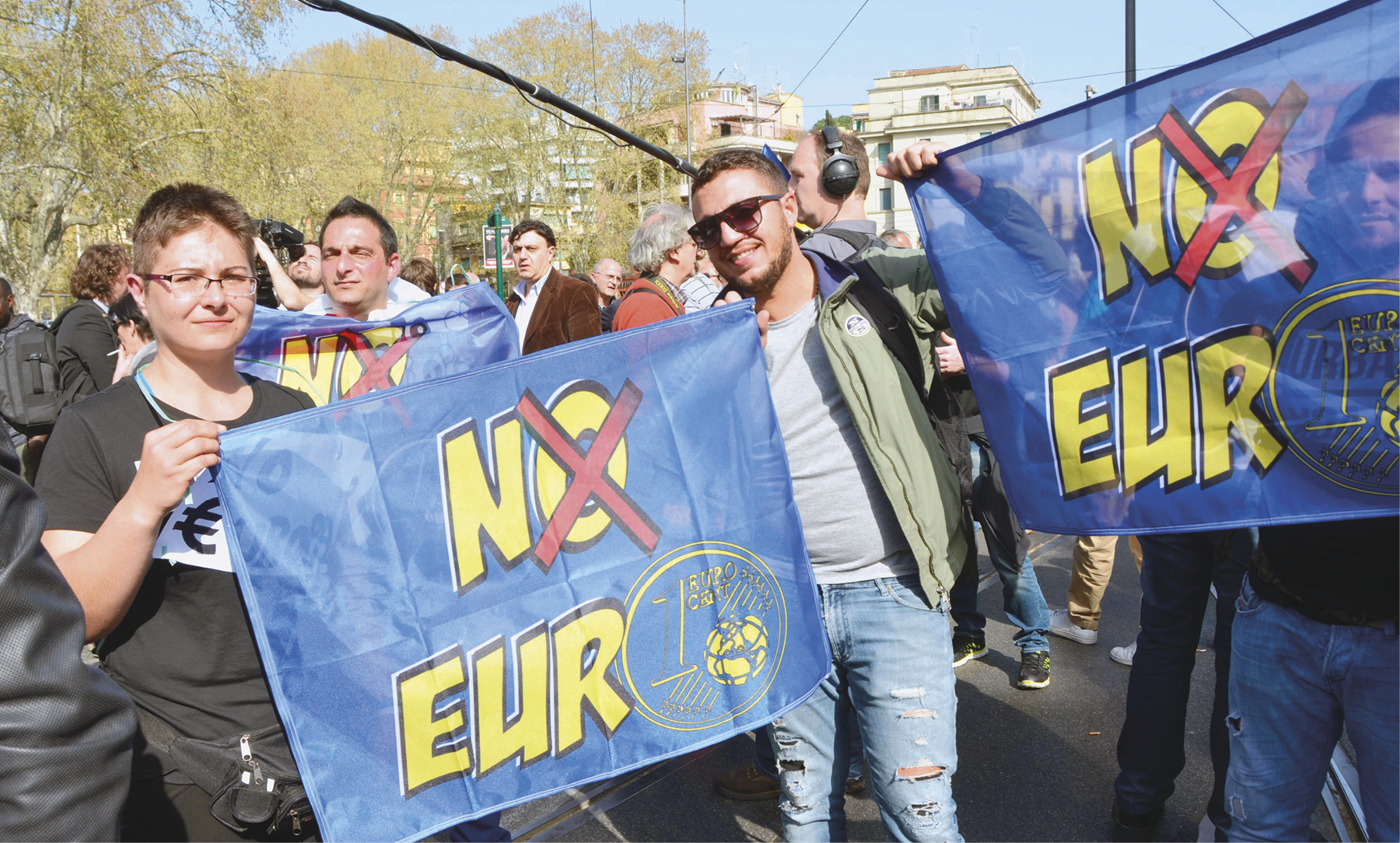 Fotografia. Manifestação de pessoas na rua, com destaque para duas pessoas segurando faixas azuis escrito: NO EURO e um xis na letra O.