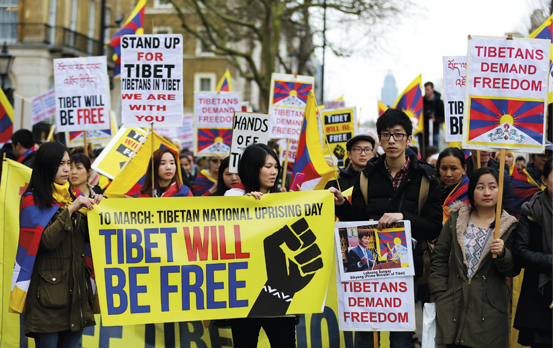 Fotografia. Manifestação de rua composta por um grupo de pessoas segurando cartazes e faixas. Destaque para faixa amarela com texto: TIBET WILL BE FREE.