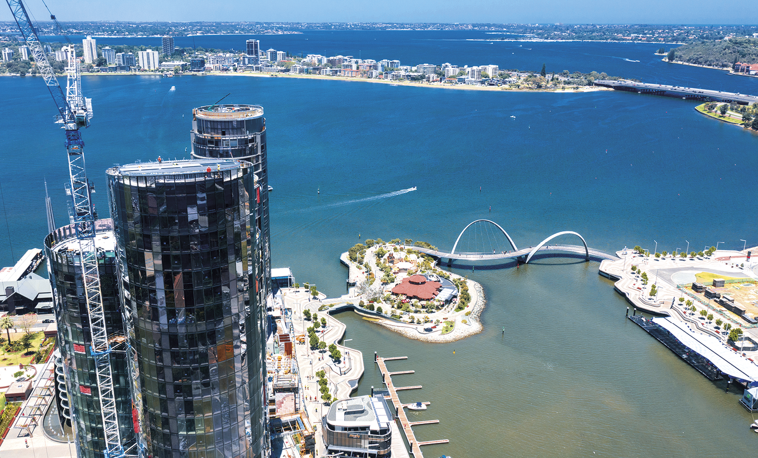 Fotografia. Vista aérea para uma cidade a beira-mar. À esquerda, arranha-céus espelhados em construção e outras edificações. À direita, um canal com uma marina e ponte ligando as duas zonas da cidade em destaque na imagem. Ao fundo, mar, ponte e outras zonas urbanas.