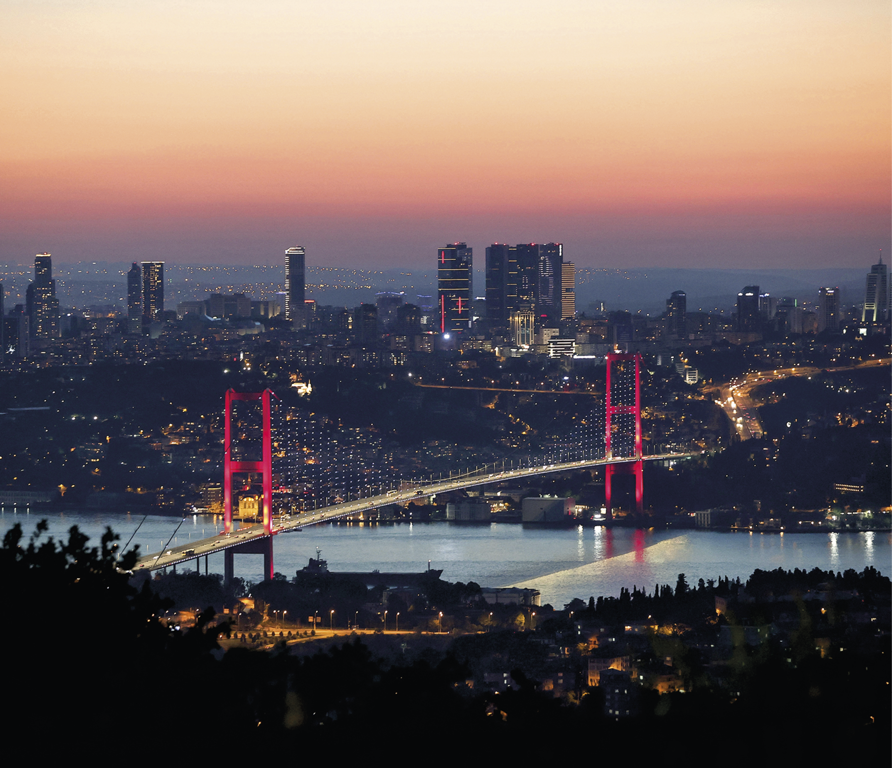 Fotografia. Vista de um curso d’água sob uma ponte pênsil iluminada de vermelho. As margens do curso d’água são ocupadas por uma densa área urbana. No plano de fundo, a área urbana é composta por edifícios altos e grande avenida que dá acesso à ponte. O céu tem cor alaranjada e há pouca luminosidade natural; as luzes das construções estão acesas.