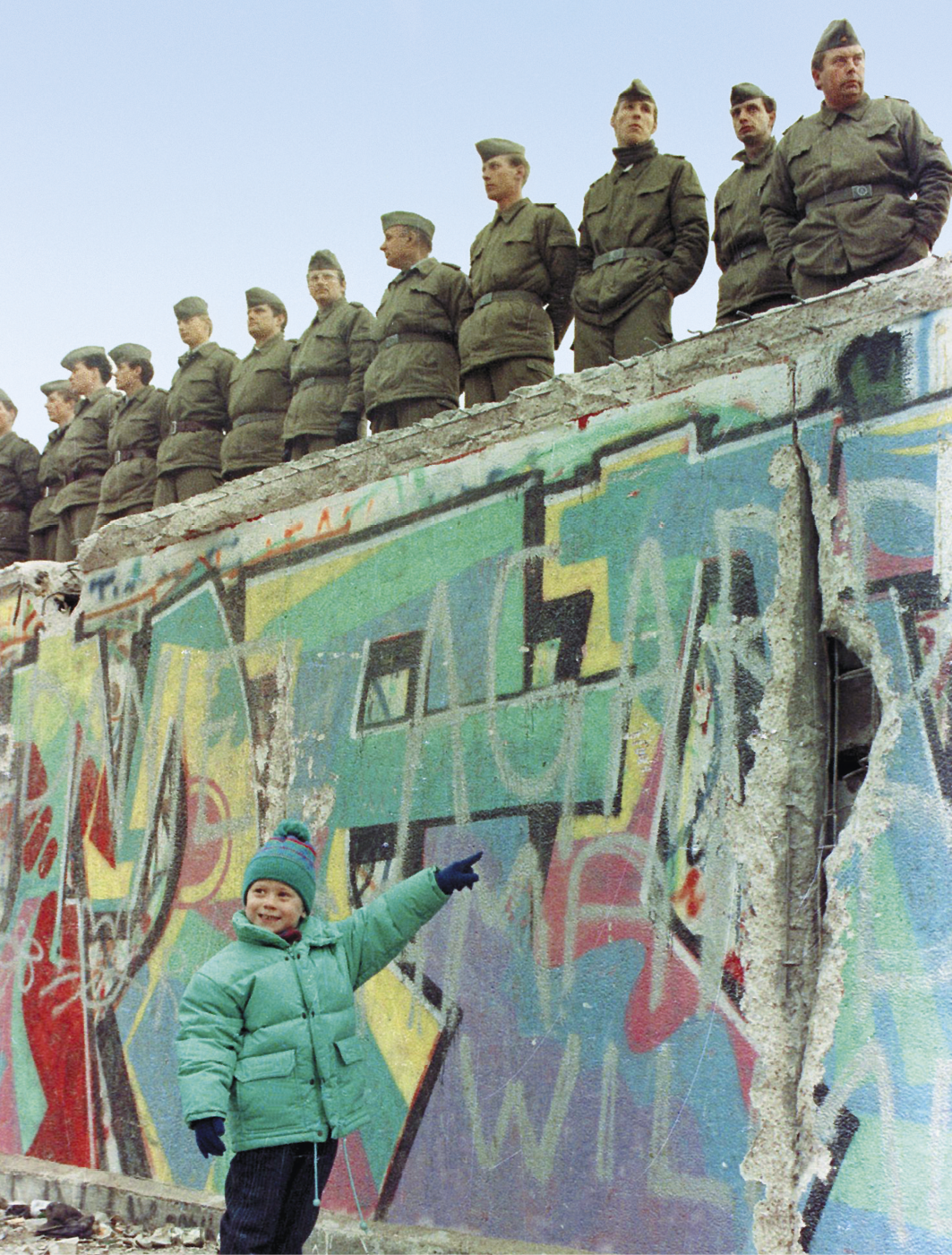 Fotografia. Destaque para uma criança de jaqueta e gorro verde, de pé, sorrindo e apontando para um muro alto, coberto de grafite colorido. Em cima do muro, há diversos soldados de uniforme militar e com as mãos para trás, posicionados um ao lado do outro.