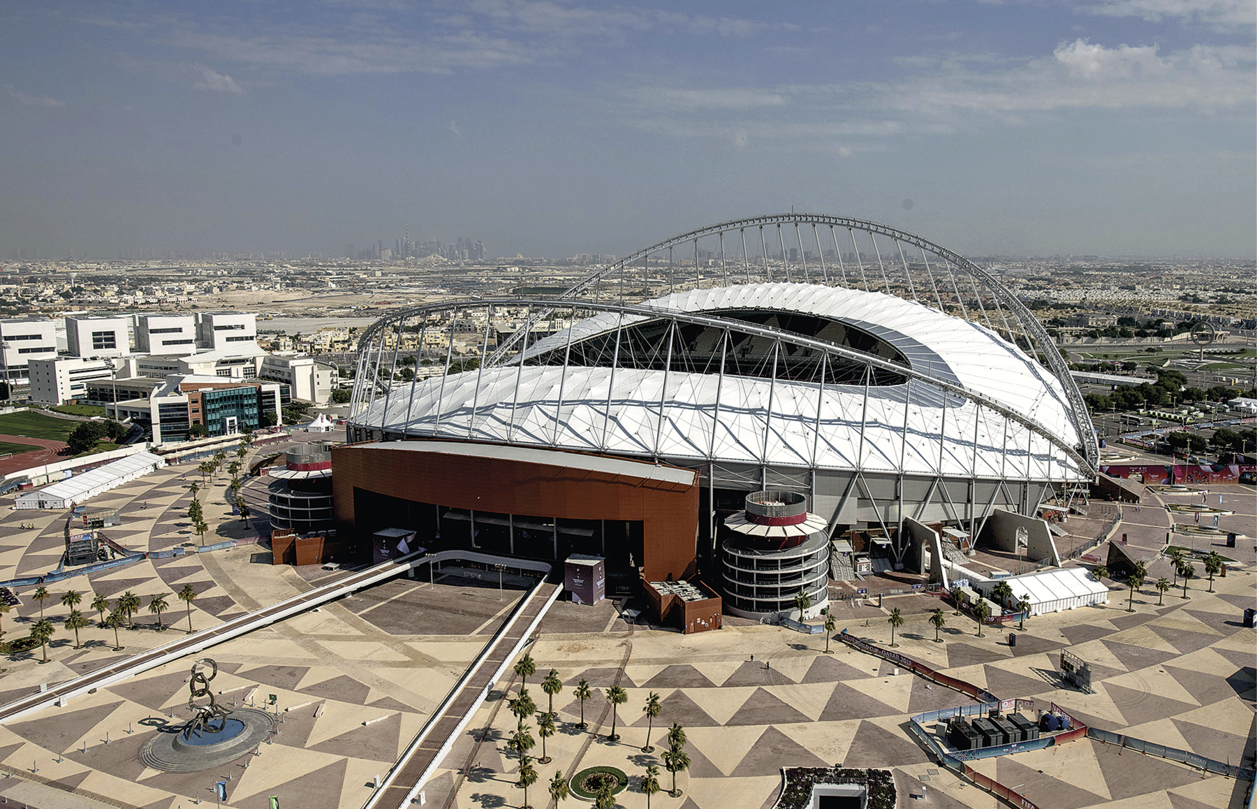 Fotografia. Vista para um estádio em formato oval, com cobertura semiaberta branca e com estruturas metálicas e prateadas. O estádio possui um portal marrom e está situado em uma superfície plana, cujo piso possui figuras triangulares. Ao redor do estádio, área urbana predominantemente horizontal.