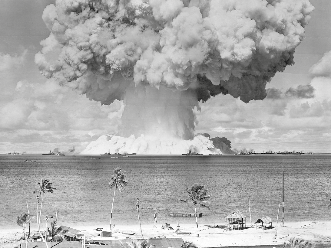 Fotografia em preto e branco. No primeiro plano, destaque para uma praia com coqueiros, quiosques e mar. No segundo plano, uma grande fumaça densa em formato de cogumelo no mar e céu com nuvens.