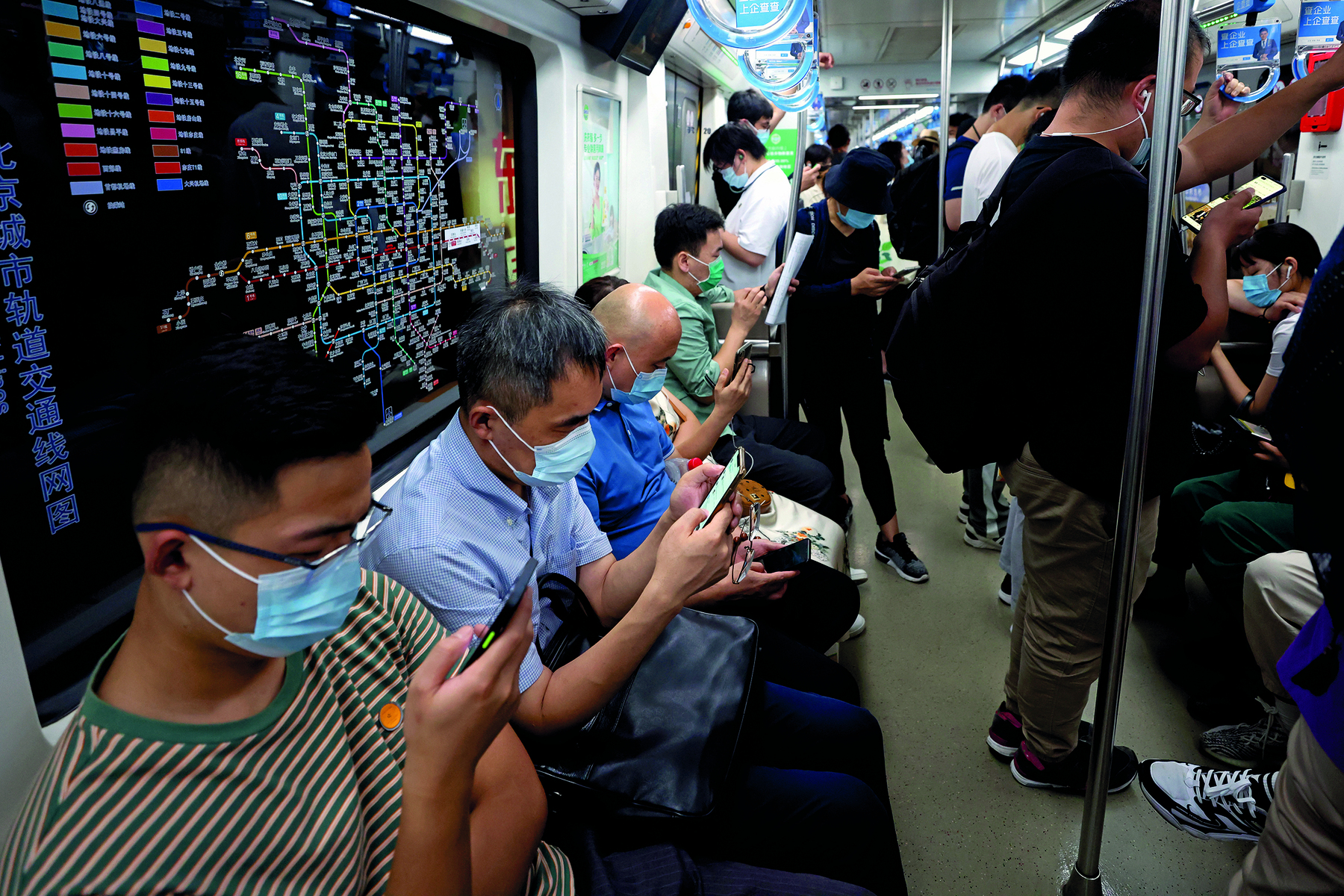 Fotografia. Vista para a área interna de um vagão de metrô. À esquerda, diversas pessoas sentadas com máscaras de proteção facial manuseiam celulares. Atrás delas, janela do trem com mapa da rede de metrô. À direita e ao fundo, diversas pessoas em pé com máscaras de proteção facial. Algumas delas manuseando o celular e com fone de ouvido.