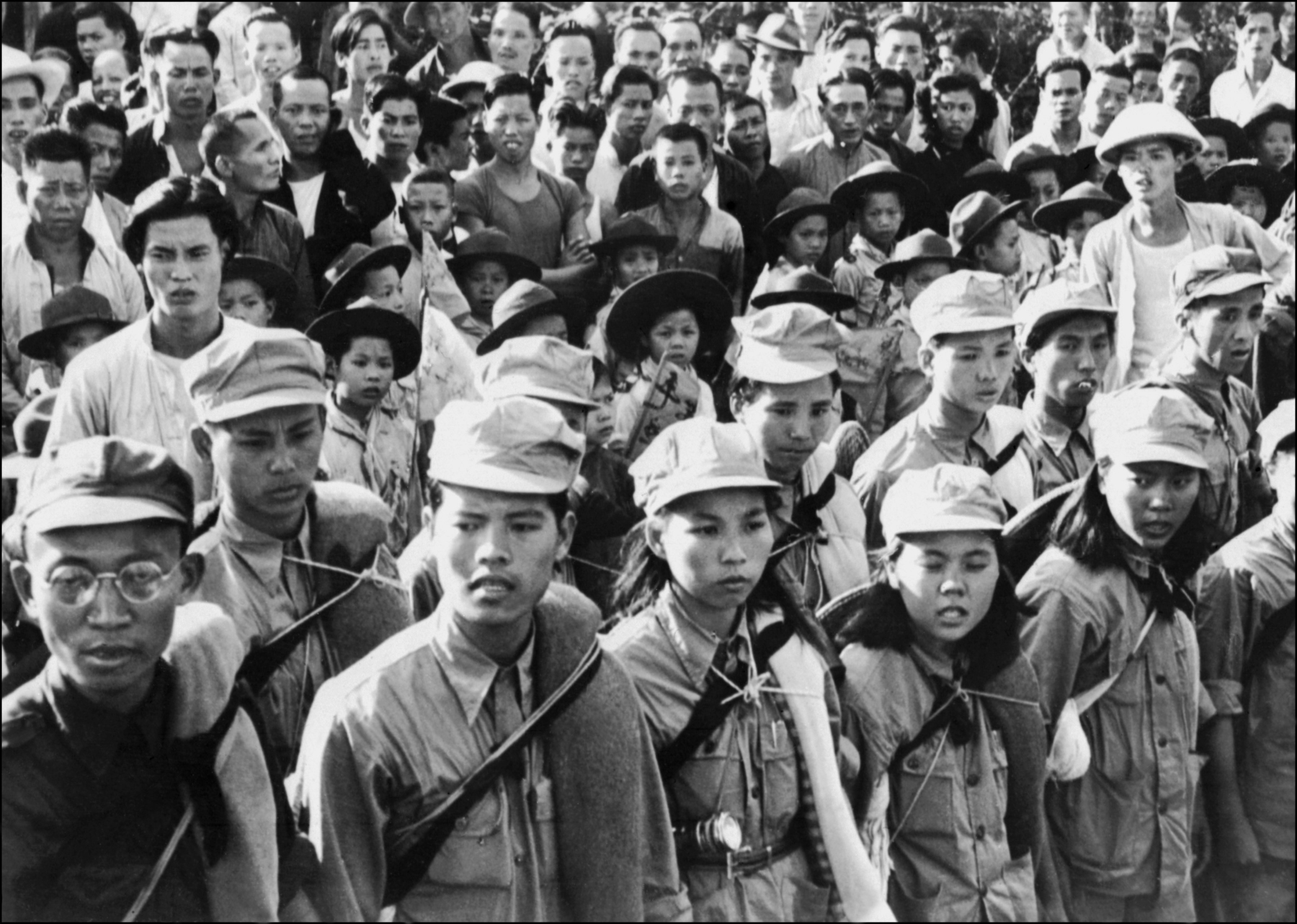 Fotografia em preto e branco. Vista para uma multidão. No primeiro plano, duas fileiras compostas por homens e mulheres vestidos com uniformes militares. Atrás, no segundo plano, diversas pessoas aglomeradas, de crianças a adultos.