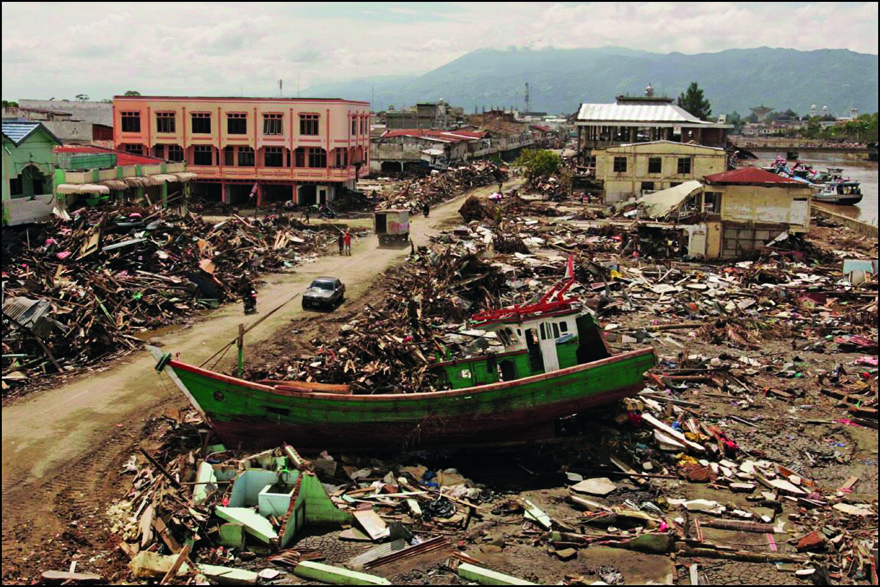 Fotografia. No primeiro plano, um barco de madeira verde tombado no chão entre entulhos e destroços que cobrem a superfície de terra de um quarteirão. Ao lado, uma rua coberta de lama seca, com dois veículos, motocicleta e quatro pessoas circulando. À esquerda, quarteirão coberto por entulhos e destroços. No segundo plano, quarteirões com entulhos e destroços entremeados de algumas edificações destruídas ou danificadas. Ao fundo, presença de montanhas e céu encoberto por nuvens.