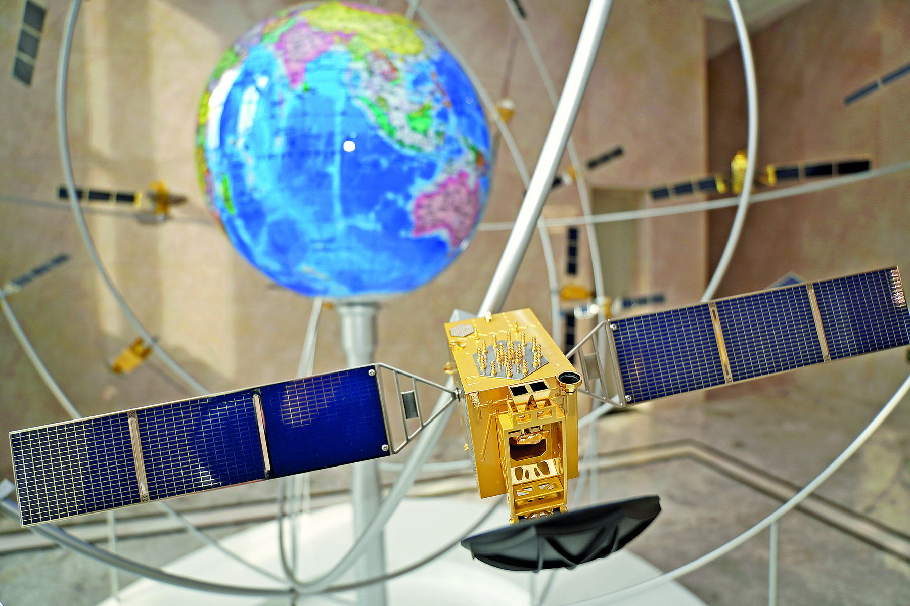 Fotografia. Vista para um salão com uma maquete. No primeiro plano, destaque para um satélite amarelo com painéis solares nas laterais. No segundo plano, o Planeta Terra disposto no meio da imagem.