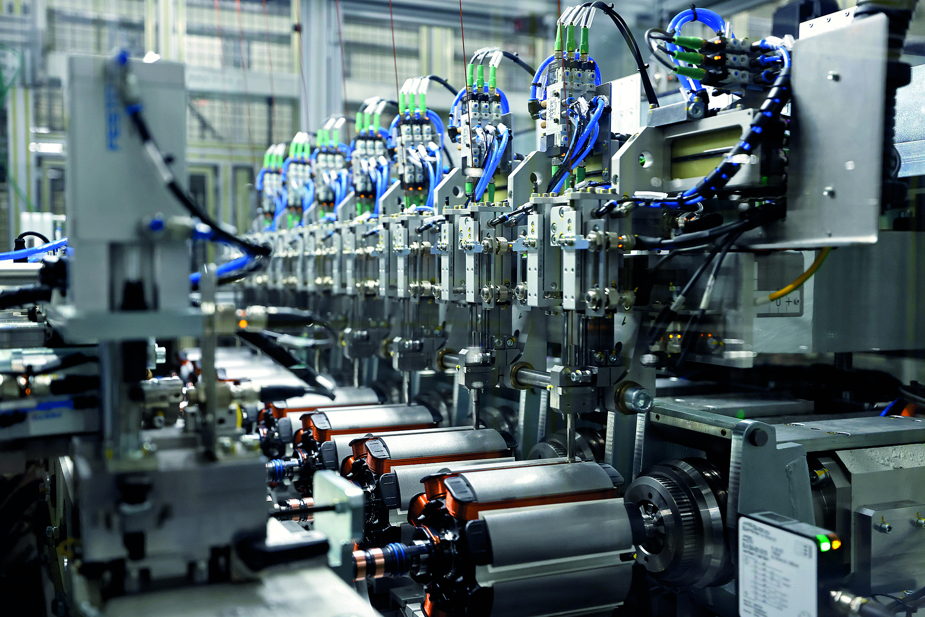 Fotografia. Destaque para uma linha de produção industrial composta apenas por máquinas cinzas com cabos azuis e pretos; na parte inferior, há mecanismos redondos.