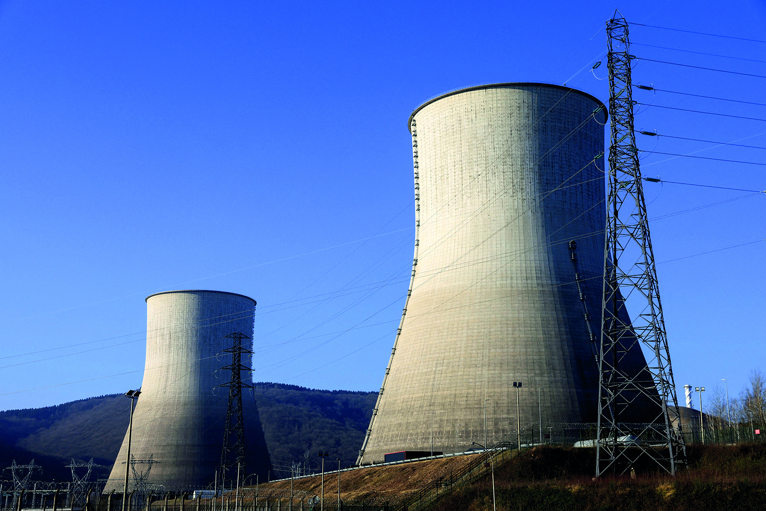 Fotografia. Destaque para duas torres de resfriamento de usinas nucleares sobre uma superfície inclinada. Ao lado direito, há uma torre de eletricidade. Ao fundo, montanha e acima, céu azul.