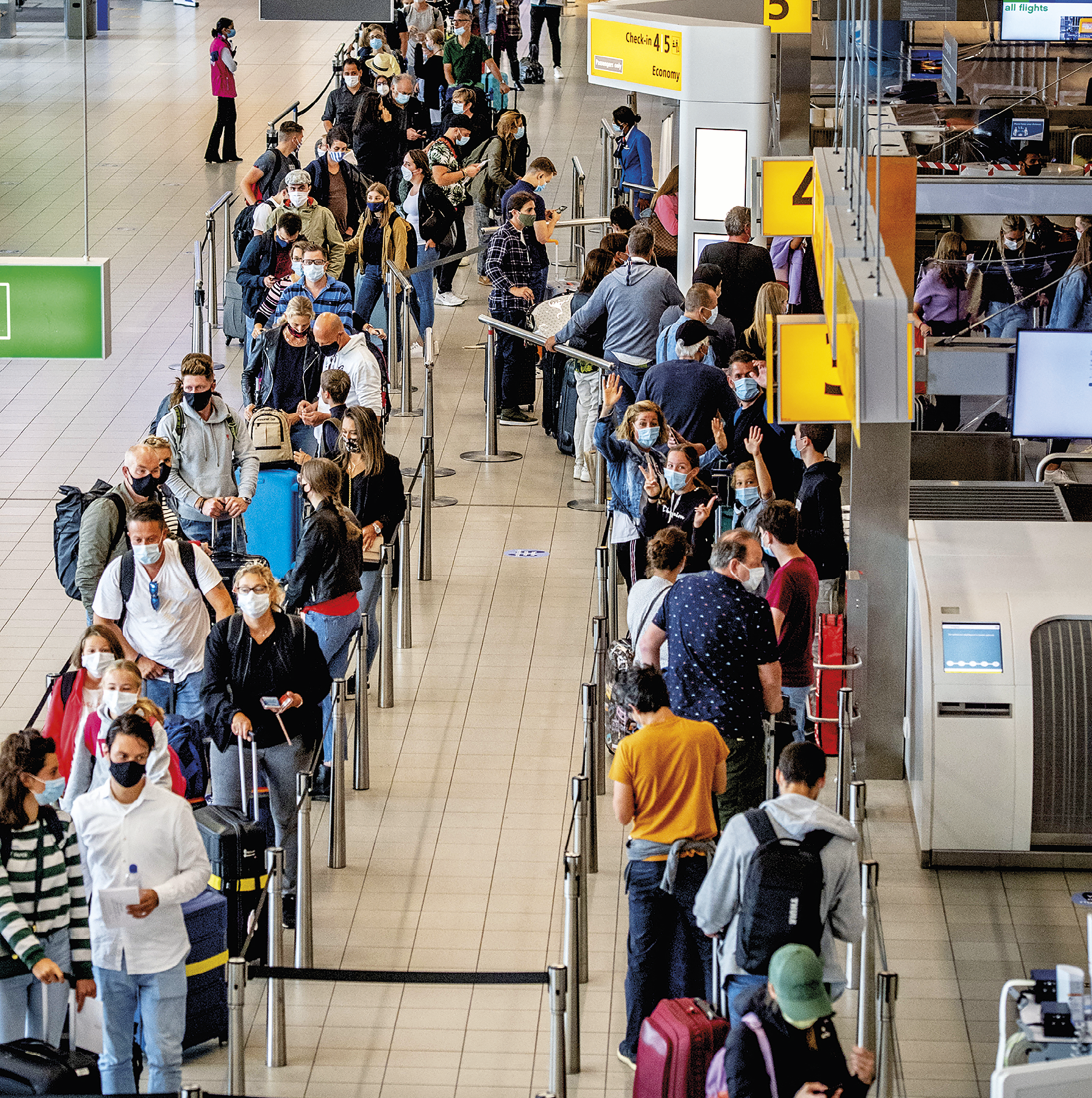 Fotografia. Vista do saguão de aeroporto. No centro, uma fila de pessoas aguardando a entrada no portão de embarque. Algumas pessoas estão de frente e outras de costas e a maior parte delas usa máscara de proteção facial e leva bagagem. Ao lado esquerdo, piso livre do saguão e uma pessoa de colete cor de rosa. Ao lado direito, guichês com atendentes e placas de sinalização amarela.