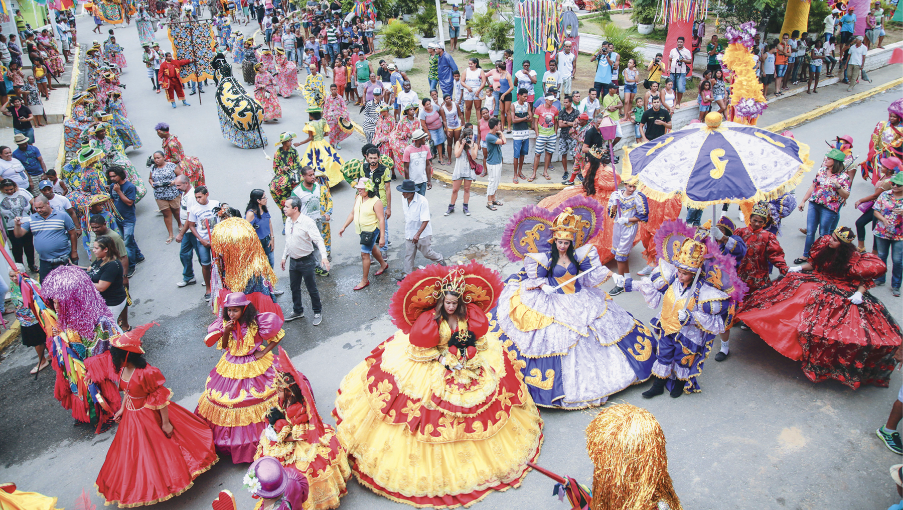 Fotografia. No primeiro plano, vista para um cruzamento de ruas ocupado por um desfile de mulheres de vestidos compridos e rodados, fantasias e adereços coloridos. No segundo plano, diversas pessoas assistem ao desfile nas calçadas.