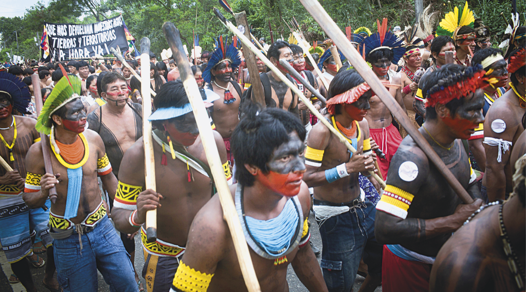 Fotografia. Destaque para uma manifestação de pessoas indígenas com o corpo e o rosto pintados, usando colares, cocares e pulseiras coloridos. Muitos deles seguram um bastão queimado na ponta e faixa. Ao fundo, há árvores.