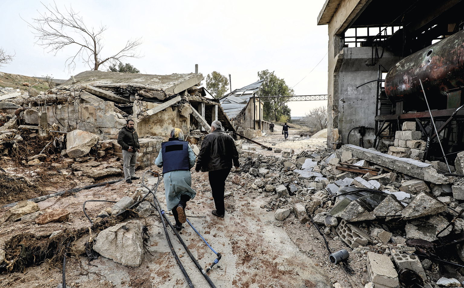 Fotografia. No centro, duas pessoas usando agasalhos estão caminhando de costas. Ao redor, casas e galpões destruídos. Abaixo, superfície com diversos fragmentos de rochas e entulhos.