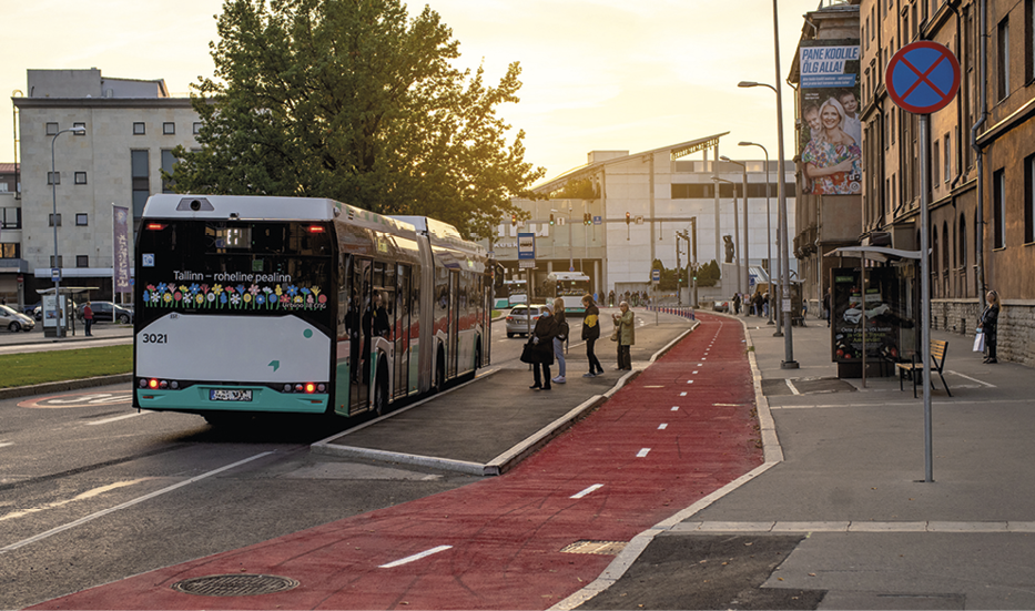 Fotografia. No centro, vista para uma ciclovia vermelha entre duas calçadas. Na calçada direita, placa de trânsito e fachada de edifício. Na calçada esquerda, pessoas em pé no ponto de ônibus. Ao lado esquerdo, rua com ônibus estacionado no ponto.