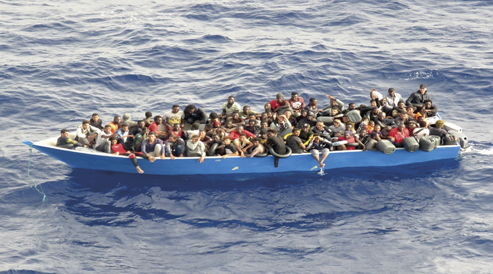 Fotografia. Destaque para um barco azul repleto de pessoas. Ao redor, o mar.