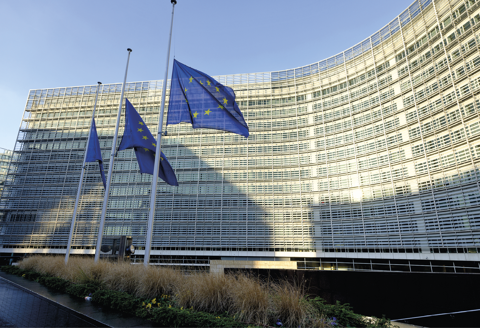 Fotografia. No primeiro plano, destaque para três mastros com as bandeiras da União Europeia sobre um pequeno jardim. Ao fundo, fachada de vidro do edifício-sede da Comissão Europeia.