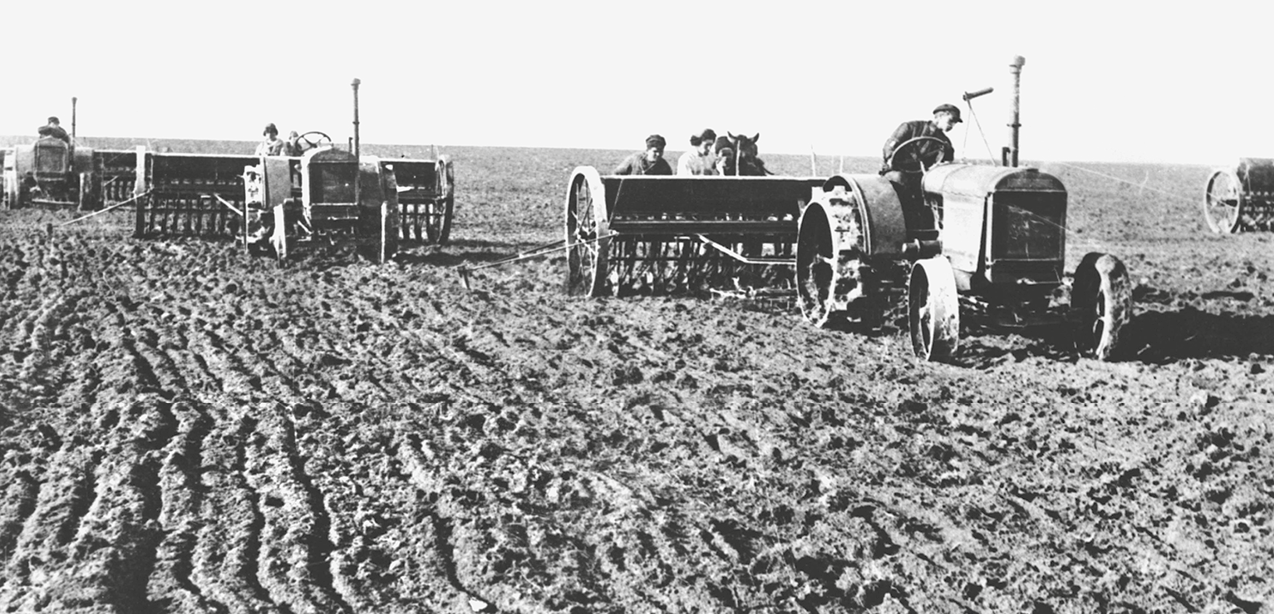 Fotografia em preto e branco. Vista para uma superfície plana ocupada por terreno, cujo solo está sendo arado por máquinas agrícolas pilotadas por pessoas.