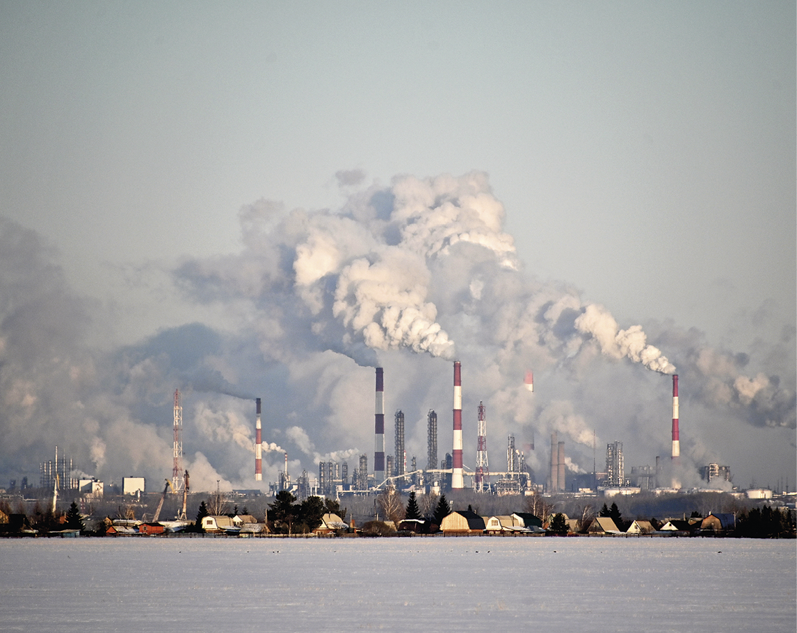 Fotografia. No primeiro plano, superfície plana sem cobertura vegetal. No segundo plano, diversos galpões, estruturas industriais e chaminés lançando fumaça na atmosfera, além de casas e árvores isoladas.