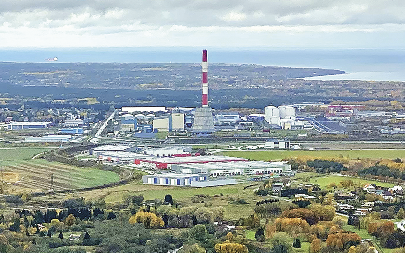 Fotografia. Vista aérea para uma superfície plana ocupada por uma zona industrial. No primeiro plano, fragmento florestal. No segundo plano, diversos galpões, construções industriais e torre alta. No terceiro plano, superfície plana e mar.