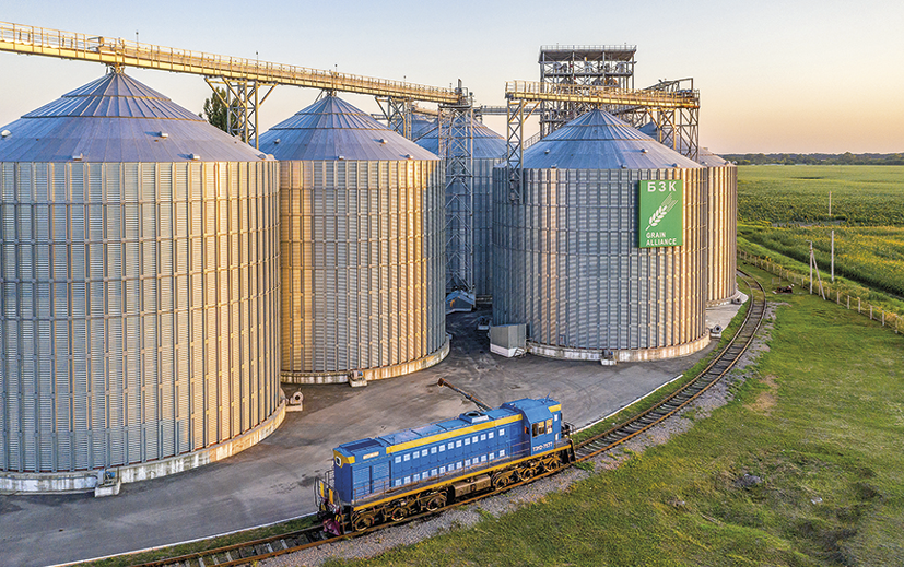 Fotografia. À direita, superfície plana coberta por vegetação rasteira. À esquerda, grandes silos arredondados e de metal e estruturas industriais. Na frente dos silos, ferrovia e trem em circulação.