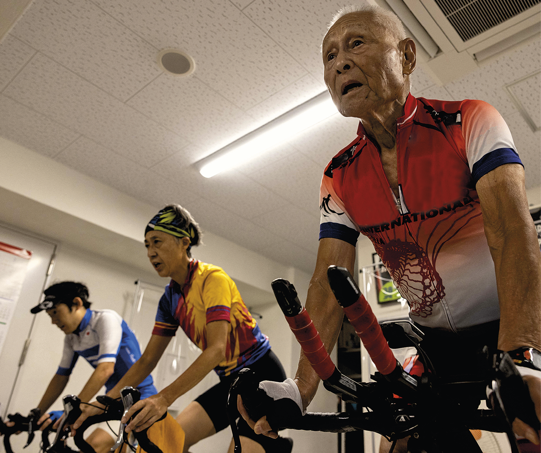 Fotografia. Vista do espaço interno de uma sala com três pessoas, duas delas idosas, pedalando em bicicletas ergométricas. As pessoas vestem roupas esportivas coloridas.