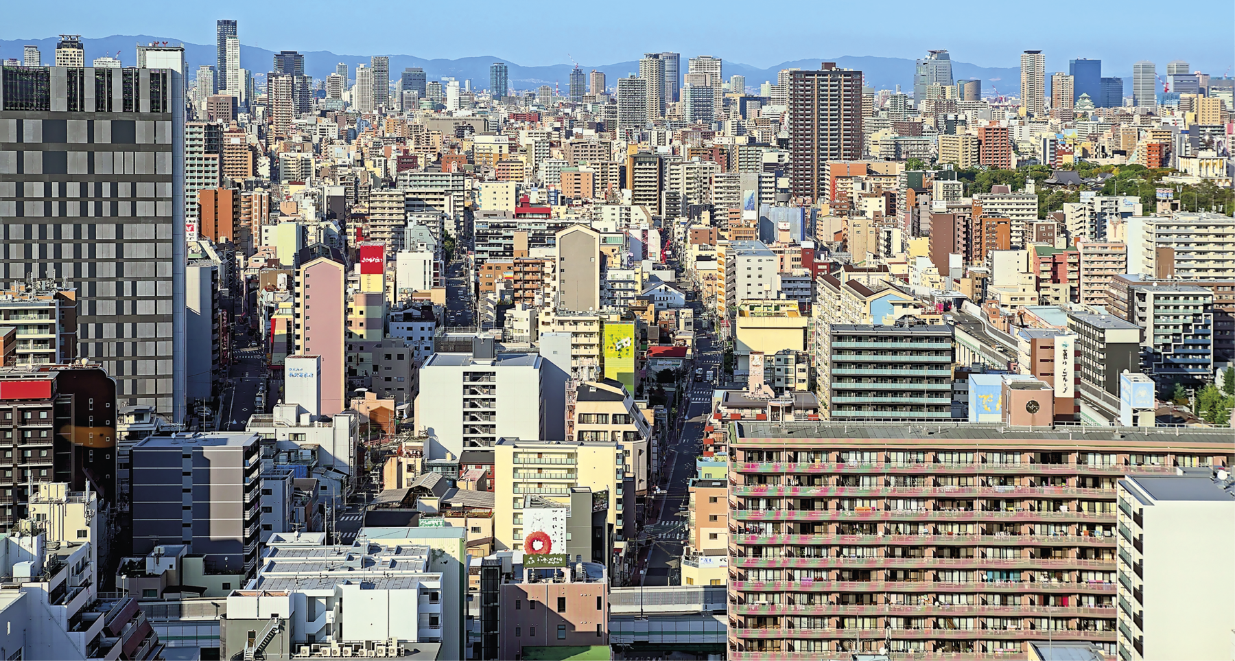 Fotografia. Vista para uma área urbana bastante adensada. No centro, uma avenida e ao redor dela, muitas edificações com estilos arquitetônicos e alturas variadas.  Ao fundo, na parte de cima da fotografia, vista de uma cadeia de montanhas e o céu azul.