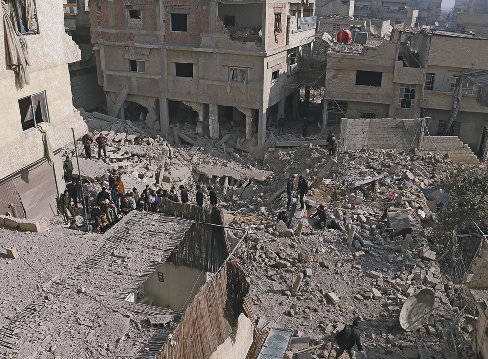 Fotografia. No centro, vista para uma superfície plana coberta por destroços de uma construção em ruínas e algumas pessoas circulando pelos entulhos. Ao redor, alguns edifícios e casas destruídos.