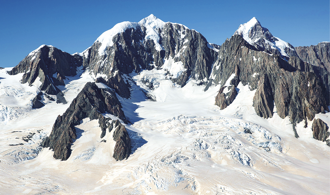 Fotografia. Vista para uma formação montanhosa com rocha exposta e algumas partes cobertas de neve.  Acima, o céu azul.