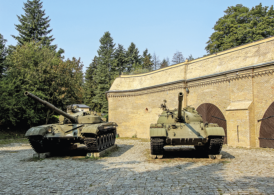 Fotografia. No primeiro plano, dois tanques de guerra verdes sobre uma superfície plana. No segundo plano, muro, árvores e céu azul.