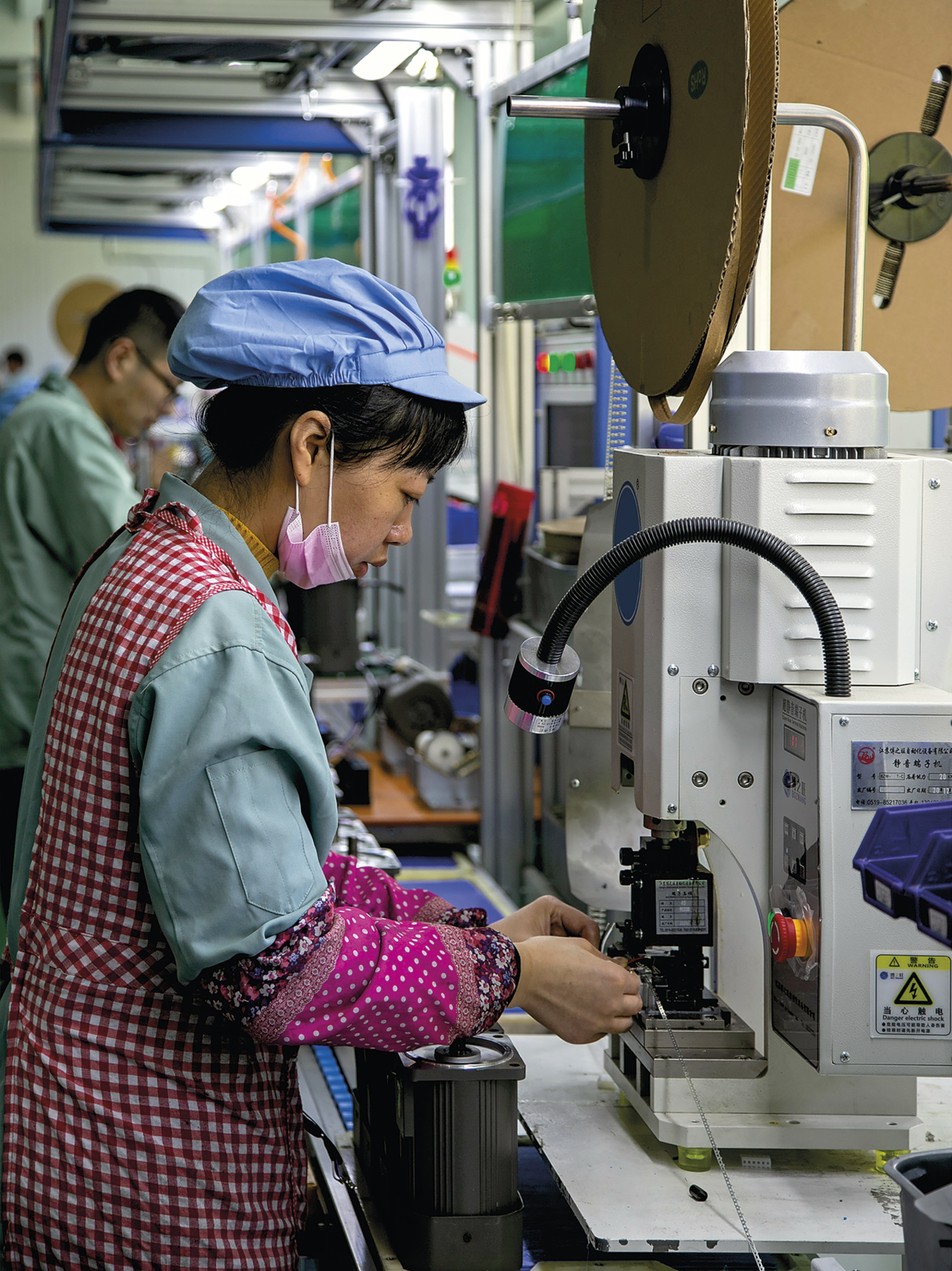 Fotografia. No primeiro plano, vista para uma linha de montagem industrial, com destaque para uma mulher de pé, usando um uniforme verde, com touca azul e avental vermelho, manuseando uma máquina apoiada sobre uma bancada. No segundo plano, ao fundo, outra pessoa de frente para a bancada, manuseando outra máquina.