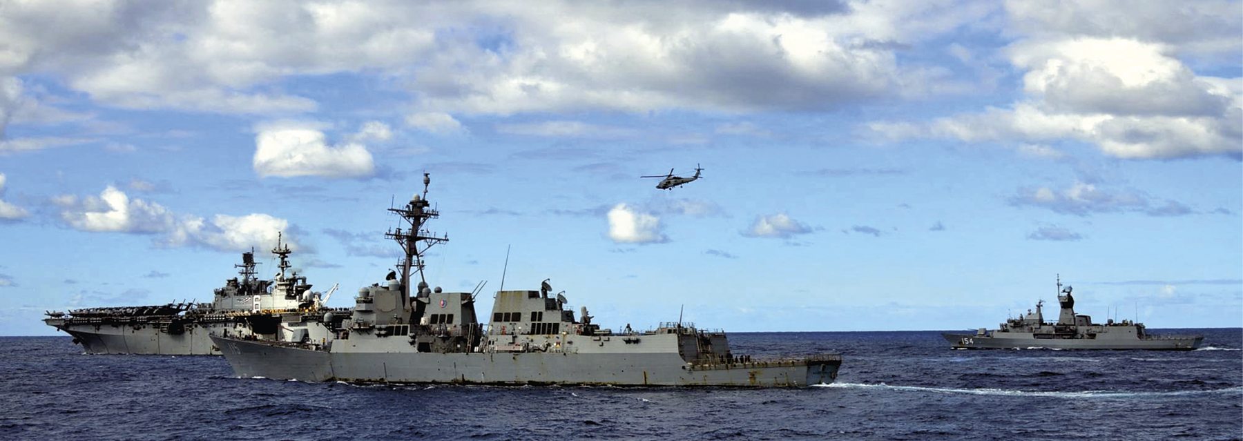 Fotografia. Vista do mar com destaque para três embarcações militares em tons de cinza com janelas e torres com antenas. Acima, o céu azul com nuvens e um helicóptero sobrevoando próximo às embarcações.