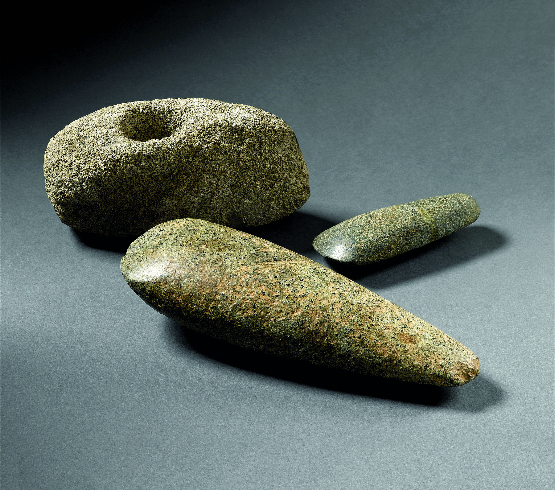 Fotografia. Três objetos de pedra em formatos e tamanhos diferentes, de cor cinza. Dois são longitudinais e bem polidos nas bordas. O outro é arredondado, e tem um furo no meio.