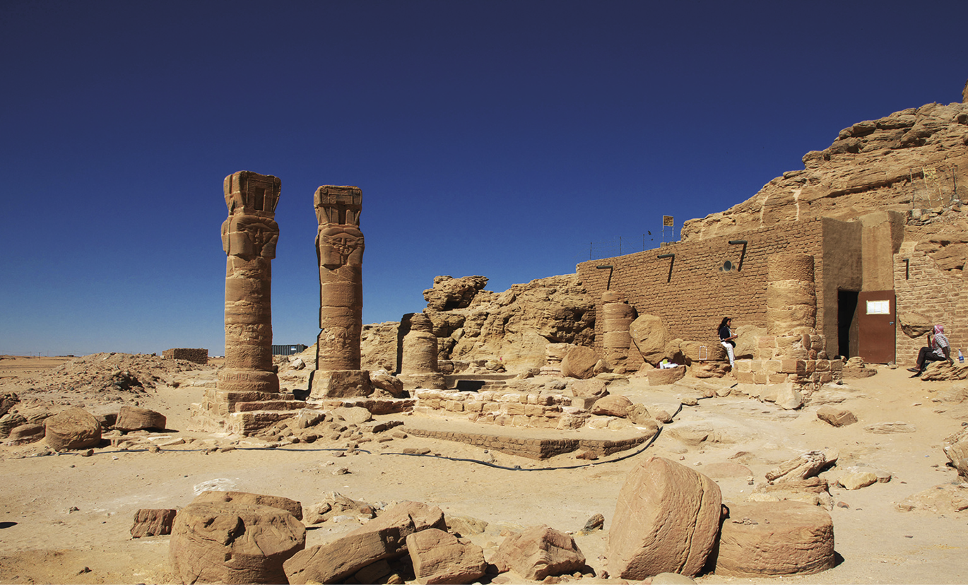 Fotografia. Sob um céu azul sem nuvens, vista de local aberto, com ruínas de construções sobre o solo arenoso, com pedras espalhadas. À esquerda, destacam-se duas colunas na vertical arredondadas e, à direita, um muro de pedras cor de areia.