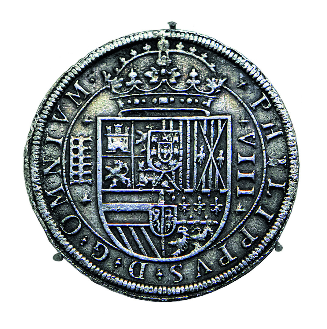 Fotografia. Uma moeda redonda na cor prata com figuras em relevo. Na parte superior, há uma coroa e, na parte inferior, um brasão. Entre a borda e o relevo do brasão, inscrições em latim.