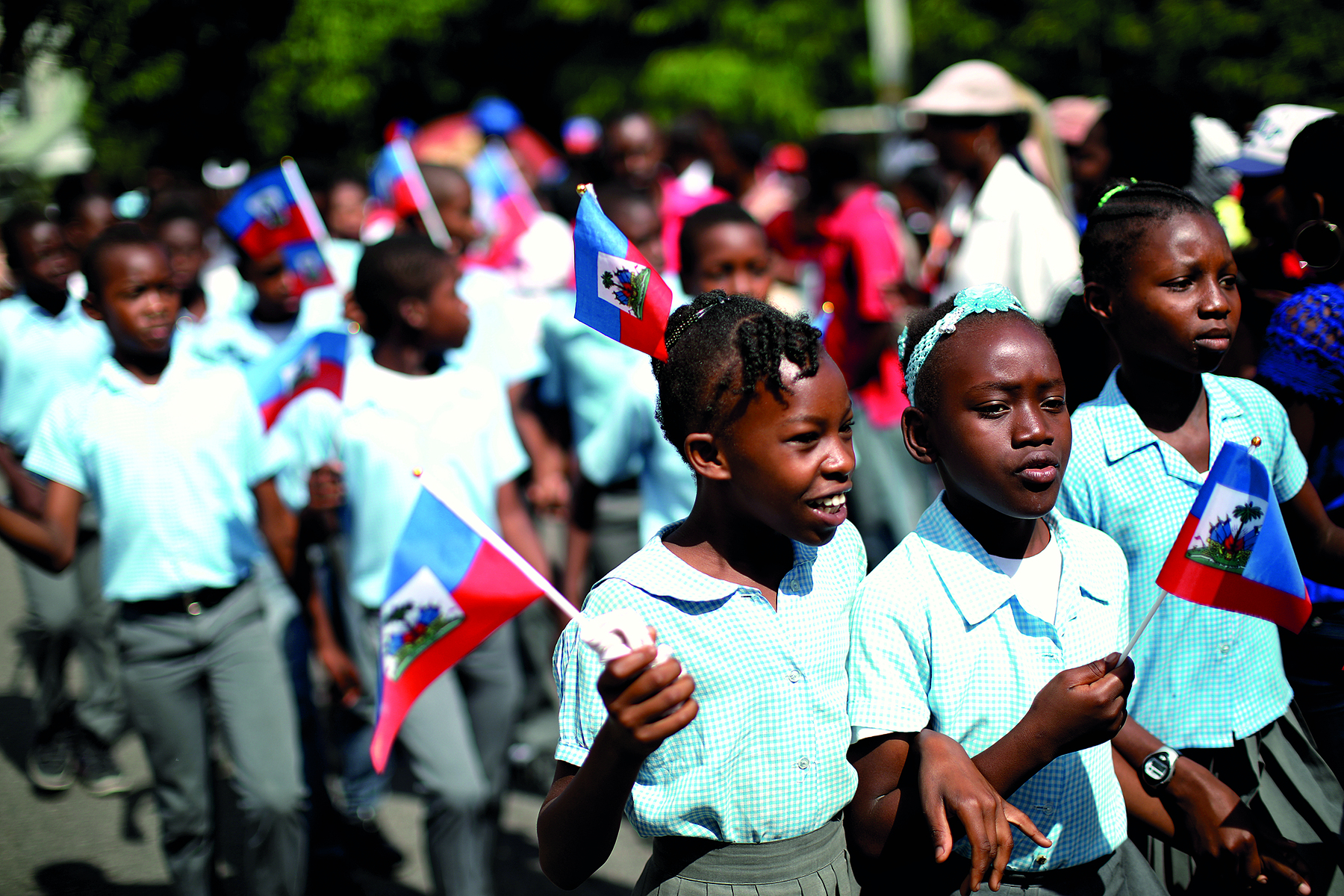 Fotografia. Dezenas de crianças em um desfile em uma rua, meninos e meninas negros de cabelos escuros, as meninas com os cabelos presos em tranças. Meninos e meninas com camisa de mangas curtas na cor azul clara, as meninas de saia de pregas de cor cinza e os meninos de calça cinza. 
À frente, três meninas juntas, duas delas de braços dados, vistas da cintura para cima. Duas delas, à esquerda, segurando pequenas bandeiras do Haiti. A menina à esquerda olha para frente, sorrindo. 
Em segundo plano, o fundo da imagem está desfocado. Há mais crianças com bandeiras e,  mais ao fundo, árvores com folhas verdes.