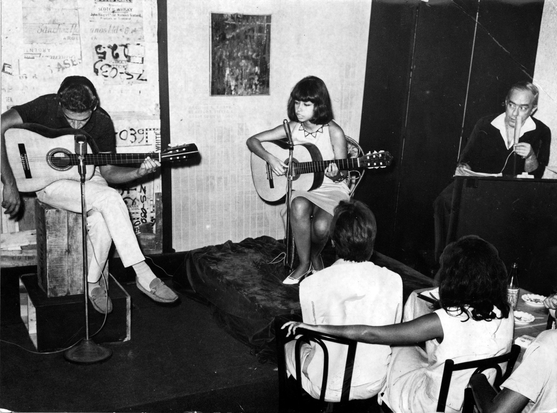 Fotografia em preto e branco. Em ambiente fechado, um homem e uma mulher, sentados, tocam violão e outro homem canta, também sentado, diante de um microfone. Na parte inferior direita, um casal sentado ao lado de uma mesa, visto de costas, observa o trio.